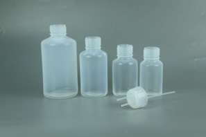 Four translucent plastic bottles of varying sizes are lined up against a plain background, with one cap and dropper placed in front.