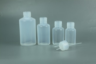 Four translucent plastic bottles of varying sizes are lined up against a plain background, with one cap and dropper placed in front.