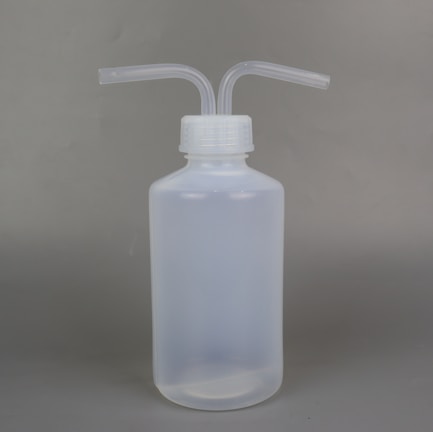 A semi-transparent plastic wash bottle with two bent dispensing nozzles. The bottle has a cylindrical shape with a white cap, and the background is a plain, neutral grey.