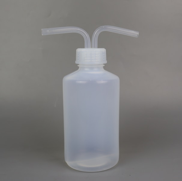 A semi-transparent plastic wash bottle with two bent dispensing nozzles. The bottle has a cylindrical shape with a white cap, and the background is a plain, neutral grey.
