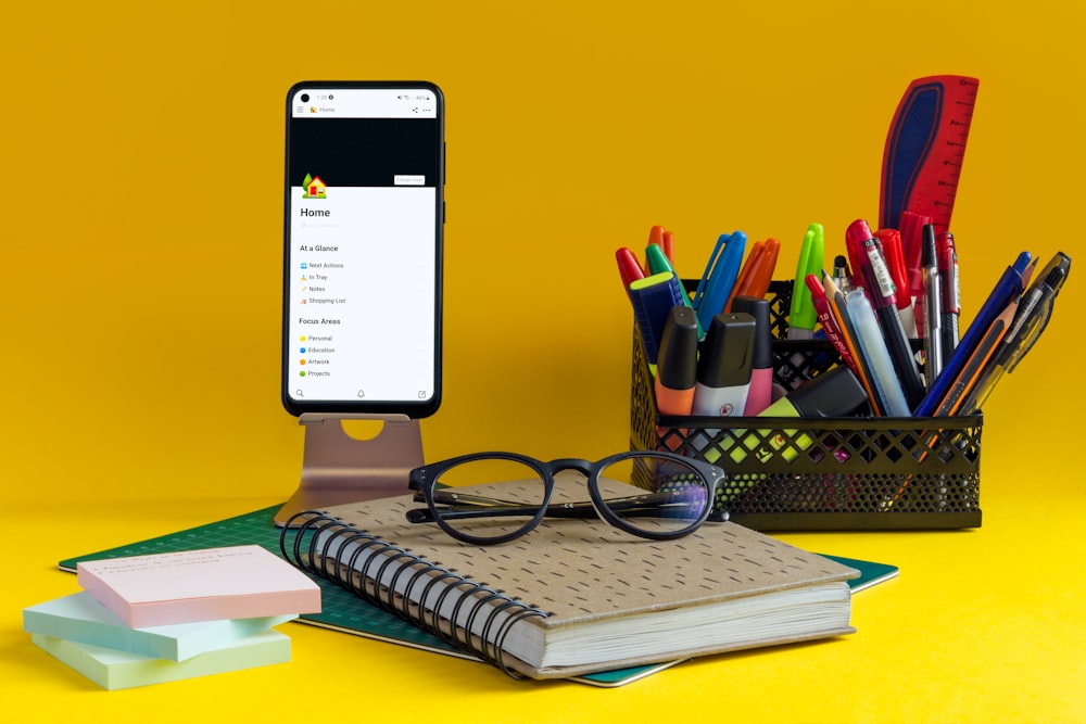 un cuaderno, bolígrafo, gafas y teléfono celular sobre un fondo amarillo