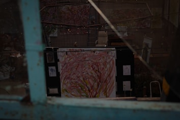 A dimly lit art studio with several abstract paintings displayed on the walls and easels. One prominent painting in the foreground features swirling strokes of pink, yellow, and red. There is a chair and some other art supplies scattered around the studio, creating an atmosphere of creativity and work in progress.