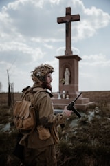A person dressed in military attire stands in front of a large cross statue in an outdoor setting. The figure holds a radio transceiver and wears a helmet with foliage attached. The background consists of a cloudy sky and open field with some grass.