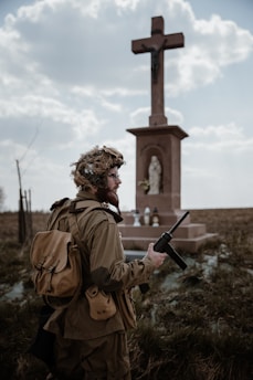 A person dressed in military attire stands in front of a large cross statue in an outdoor setting. The figure holds a radio transceiver and wears a helmet with foliage attached. The background consists of a cloudy sky and open field with some grass.