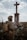 A person dressed in military attire stands in front of a large cross statue in an outdoor setting. The figure holds a radio transceiver and wears a helmet with foliage attached. The background consists of a cloudy sky and open field with some grass.