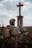 A person dressed in military attire stands in front of a large cross statue in an outdoor setting. The figure holds a radio transceiver and wears a helmet with foliage attached. The background consists of a cloudy sky and open field with some grass.