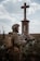 A person dressed in military attire stands in front of a large cross statue in an outdoor setting. The figure holds a radio transceiver and wears a helmet with foliage attached. The background consists of a cloudy sky and open field with some grass.