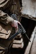 A person wearing a brownish military uniform carrying a submachine gun is depicted in an outdoor setting, standing in front of a wooden structure with blurred background.