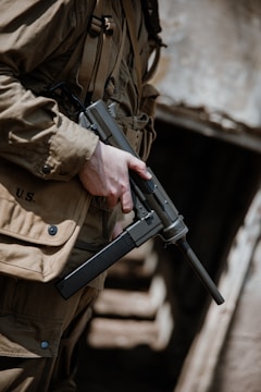 A person wearing a brownish military uniform carrying a submachine gun is depicted in an outdoor setting, standing in front of a wooden structure with blurred background.