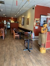 A cozy restaurant interior with wooden flooring and neatly arranged tables and chairs. A statue of a traditional figure is positioned near the entrance. Wall decorations include tapestries and framed artwork. The lighting is soft and ambient, creating a warm atmosphere.