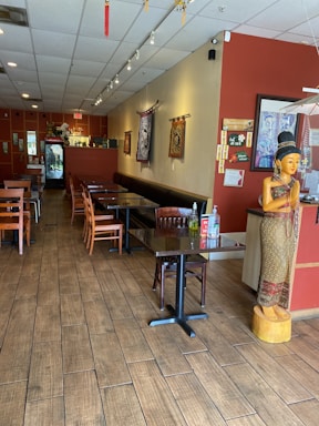 A cozy restaurant interior with wooden flooring and neatly arranged tables and chairs. A statue of a traditional figure is positioned near the entrance. Wall decorations include tapestries and framed artwork. The lighting is soft and ambient, creating a warm atmosphere.