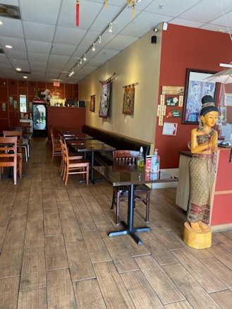 A cozy restaurant interior with wooden flooring and neatly arranged tables and chairs. A statue of a traditional figure is positioned near the entrance. Wall decorations include tapestries and framed artwork. The lighting is soft and ambient, creating a warm atmosphere.