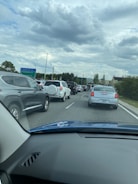 Sparse traffic is seen on a multi-lane road surrounded by greenery and illuminated traffic lights under a cloudy sky.