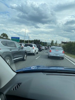 Sparse traffic is seen on a multi-lane road surrounded by greenery and illuminated traffic lights under a cloudy sky.