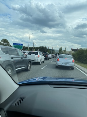 Sparse traffic is seen on a multi-lane road surrounded by greenery and illuminated traffic lights under a cloudy sky.