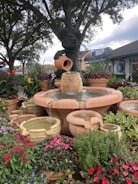 A beautifully landscaped garden features a central fountain with cascading water from several terracotta pots, surrounded by lush greenery and vibrant flowers. In the background, a decorative topiary in the shape of a character is visible, set against mature trees and a serene outdoor setting.