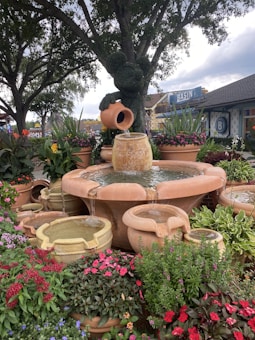 A beautifully landscaped garden features a central fountain with cascading water from several terracotta pots, surrounded by lush greenery and vibrant flowers. In the background, a decorative topiary in the shape of a character is visible, set against mature trees and a serene outdoor setting.