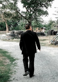 A person in a dark suit walks along a gravel path surrounded by greenery and trees. The setting appears to be semi-rural, with various tires stacked on the side and a small structure in the background. The person carries a small object in their left hand.