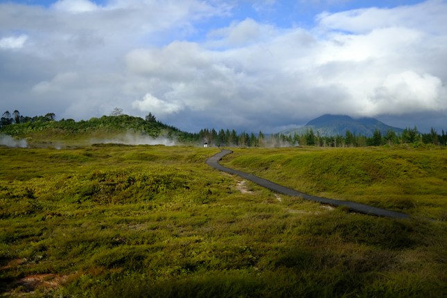 A winding path leads through lush green meadows with mist rising in the distance. In the background, a range of hills is partially obscured by clouds under a bright blue sky with patches of white clouds.