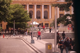 A busy outdoor scene in front of a grand, classical building labeled 'Teatro Nacional' featuring tall white columns, arched windows, and yellow walls. There is a crowd of people walking and socializing around the area. Some people are sitting near a large, lush green tree, while others are standing near steps that lead up to the building. Pigeons can be seen scattered on the ground and flying around. There are street lamps and informational signs visible in the surrounding area.