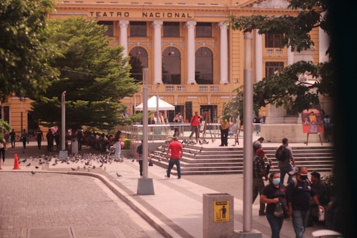 A busy outdoor scene in front of a grand, classical building labeled 'Teatro Nacional' featuring tall white columns, arched windows, and yellow walls. There is a crowd of people walking and socializing around the area. Some people are sitting near a large, lush green tree, while others are standing near steps that lead up to the building. Pigeons can be seen scattered on the ground and flying around. There are street lamps and informational signs visible in the surrounding area.