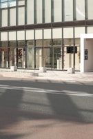 A modern building facade with large glass windows and a sleek, minimalist design. The entrance features transparent doors, and some interior elements are visible through the windows, including decorations and furniture. The exterior includes a sidewalk with stone blocks and a roadway in the foreground.
