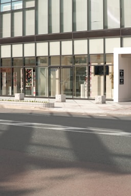 A modern building facade with large glass windows and a sleek, minimalist design. The entrance features transparent doors, and some interior elements are visible through the windows, including decorations and furniture. The exterior includes a sidewalk with stone blocks and a roadway in the foreground.