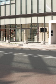 A modern building facade with large glass windows and a sleek, minimalist design. The entrance features transparent doors, and some interior elements are visible through the windows, including decorations and furniture. The exterior includes a sidewalk with stone blocks and a roadway in the foreground.