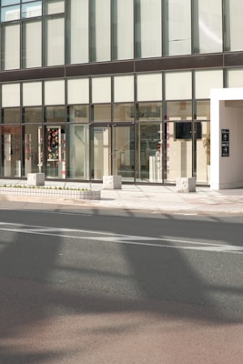 A modern building facade with large glass windows and a sleek, minimalist design. The entrance features transparent doors, and some interior elements are visible through the windows, including decorations and furniture. The exterior includes a sidewalk with stone blocks and a roadway in the foreground.