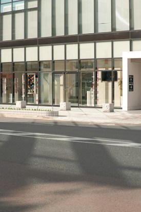 A modern building facade with large glass windows and a sleek, minimalist design. The entrance features transparent doors, and some interior elements are visible through the windows, including decorations and furniture. The exterior includes a sidewalk with stone blocks and a roadway in the foreground.