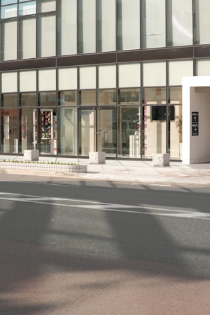 A modern building facade with large glass windows and a sleek, minimalist design. The entrance features transparent doors, and some interior elements are visible through the windows, including decorations and furniture. The exterior includes a sidewalk with stone blocks and a roadway in the foreground.