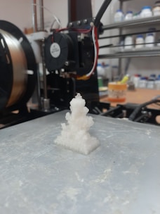 A 3D printer in a workshop setting, featuring a partially printed white object on the build plate. Behind the printer, there are shelves with various containers, suggesting a workspace. The machine is equipped with visible wires and components.