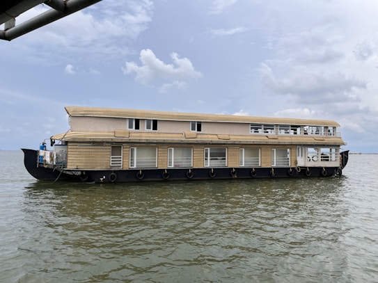 A large, two-story houseboat is floating on calm waters under a partly cloudy sky. The boat has multiple windows and a beige exterior with black trim. The water appears to be a tranquil body of water such as a lake or river.