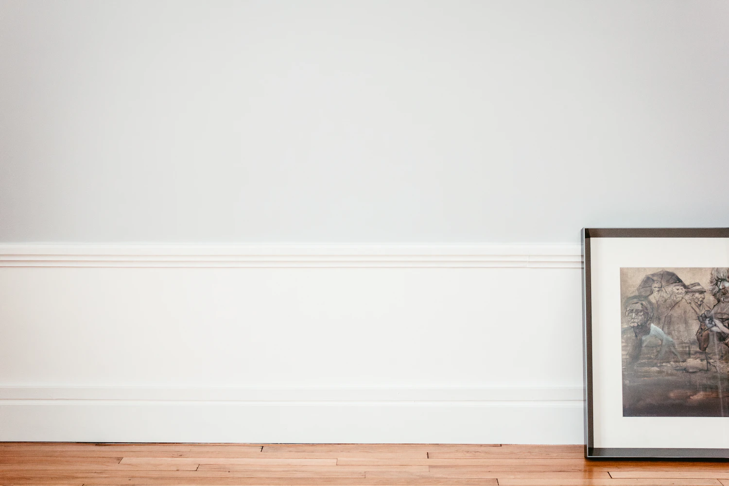 A single framed picture leaning on the skirting board of a tall, empty white wall above a warm wooden floor