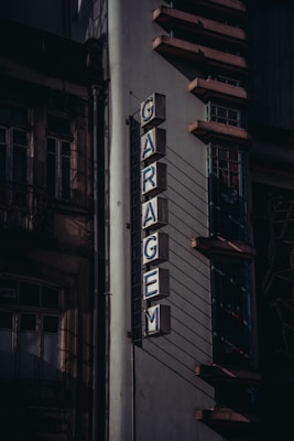 An old building facade with a vertical neon sign spelling 'GARAGEM' in block letters. The sign is mounted to the side of the building, which features architectural details such as decorative ledges and multiple windows with intricate metalwork. The lighting creates shadows that add depth and a sense of age to the structure.