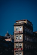 A multi-story building with a light beige façade and ornate railings, featuring red and white signs with text in Portuguese. The building stands against a clear blue sky, with other structures visible in the background.