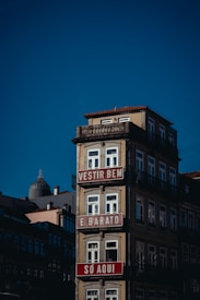 A multi-story building with a light beige fa&ccedil;ade and ornate railings, featuring red and white signs with text in Portuguese. The building stands against a clear blue sky, with other structures visible in the background.