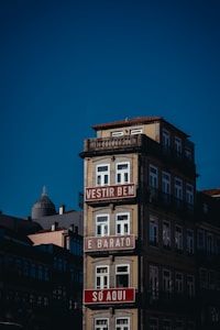 A multi-story building with a light beige fa&ccedil;ade and ornate railings, featuring red and white signs with text in Portuguese. The building stands against a clear blue sky, with other structures visible in the background.