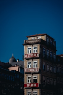 A multi-story building with a light beige façade and ornate railings, featuring red and white signs with text in Portuguese. The building stands against a clear blue sky, with other structures visible in the background.