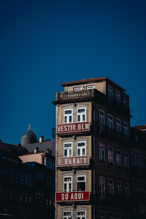A multi-story building with a light beige façade and ornate railings, featuring red and white signs with text in Portuguese. The building stands against a clear blue sky, with other structures visible in the background.