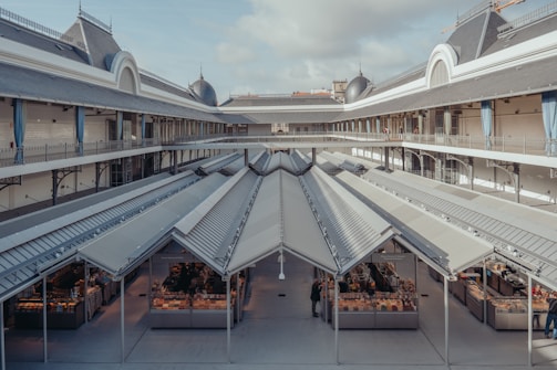 A spacious indoor market with a high roof and symmetrical design features multiple small stalls selling goods. The building has a historical architectural style with two levels, decorative railings, and soft blue curtains.