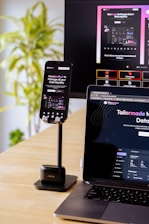 A mobile phone is held by a stand placed on a wooden table; the phone screen displays a website with financial information. A laptop beside it shows the same website, and in the background, a large monitor displays a more extensive view of the same content. A green, leafy plant is visible to the side, suggesting an indoor setting.