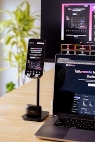 A mobile phone is held by a stand placed on a wooden table; the phone screen displays a website with financial information. A laptop beside it shows the same website, and in the background, a large monitor displays a more extensive view of the same content. A green, leafy plant is visible to the side, suggesting an indoor setting.