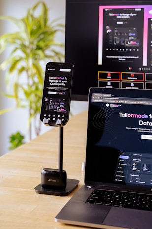 A mobile phone is held by a stand placed on a wooden table; the phone screen displays a website with financial information. A laptop beside it shows the same website, and in the background, a large monitor displays a more extensive view of the same content. A green, leafy plant is visible to the side, suggesting an indoor setting.