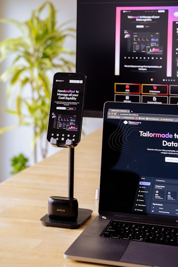 A mobile phone is held by a stand placed on a wooden table; the phone screen displays a website with financial information. A laptop beside it shows the same website, and in the background, a large monitor displays a more extensive view of the same content. A green, leafy plant is visible to the side, suggesting an indoor setting.