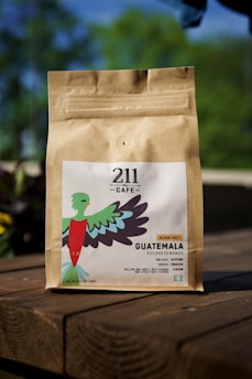 A brown paper coffee bag is sitting on a wooden table outdoors, featuring a colorful bird illustration and text detailing the coffee's origin from Guatemala and its medium roast profile.