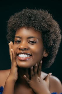 A woman with curly hair is smiling brightly, her hands gently touching her face. The background is dark, highlighting her expressive and joyful demeanor.