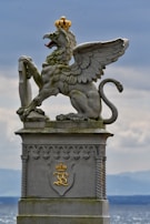 A stone statue of a mythical creature, possibly a griffin, adorned with a golden crown perched on top of its head. The creature has the body of a lion, the wings of an eagle, and is standing on a large pedestal. The pedestal features intricate carvings and a golden emblem. The background shows a cloudy sky above a distant horizon, possibly near a body of water.