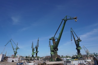 A shipyard with multiple large, industrial cranes standing tall against a clear blue sky. The cranes are green and are positioned at various angles. Below the cranes, there are several industrial buildings and equipment scattered around the yard.