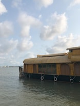 A traditional houseboat floats on a calm body of water with a partly cloudy sky overhead. The structure has a thatched roof and several circular windows, designed for cozy accommodation. In the distance, a shoreline with buildings is visible.